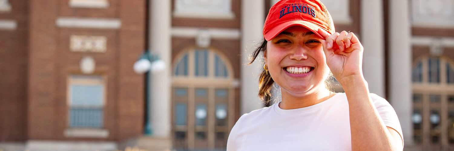 student smiling and wearing a UIUC hat on the Quad in front of Foellinger Auditorium