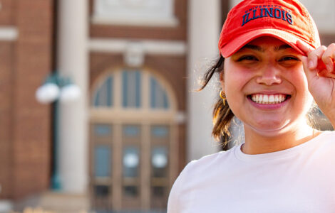 student smiling and wearing a UIUC hat on the Quad in front of Foellinger Auditorium