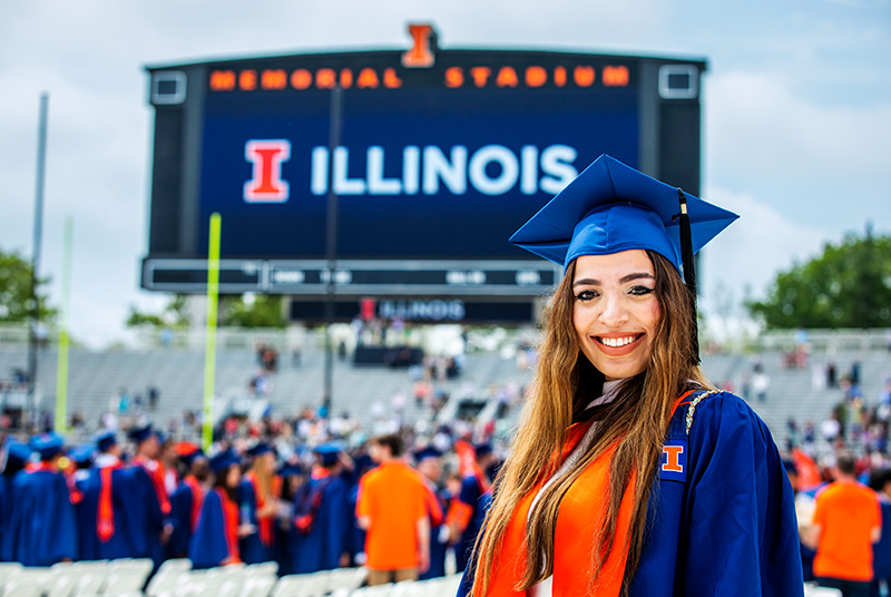 graduating student in cap and gown poses at Commencement at Memorial Stadium with the jumbotron in the background proudly displaying the orange and blue Illinois logo
