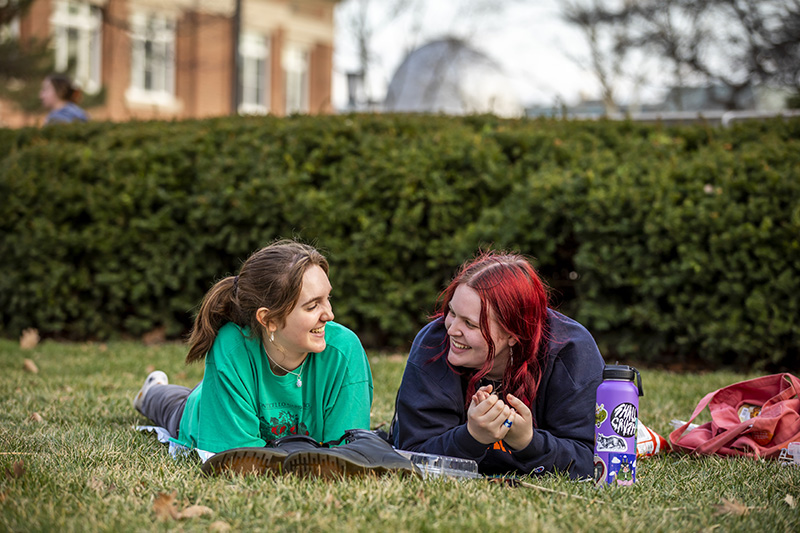 Two friends smiling and laughing together while laying on the Quad
