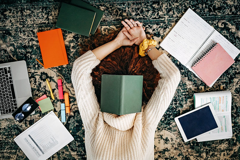 student at home laying on the floor with her face covered by a book surrounded by her laptop, notebooks, and a variety of work